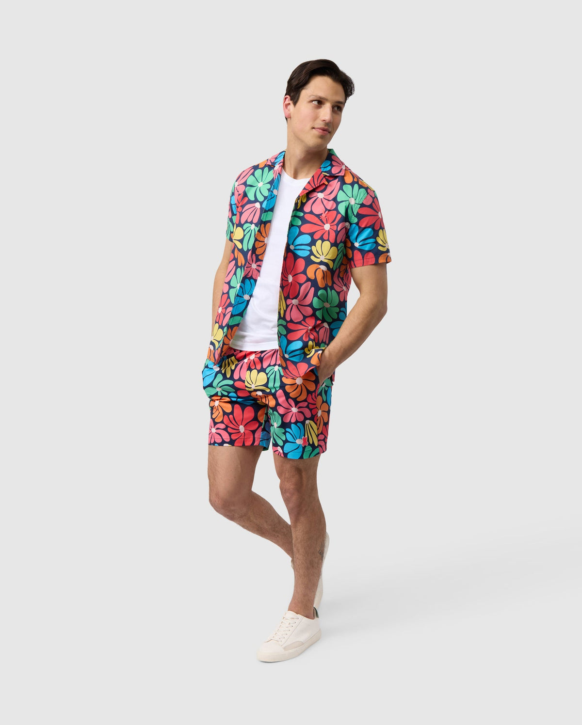 A man stands against a plain background, wearing brightly colored floral pattern swimwear with matching shorts. He has a white T-shirt underneath and is sporting white sneakers. His hands are in his Psycho Bunny MENS MENTZ SWIM TRUNK - B6W320B2SW pockets, and he gazes off to the side.