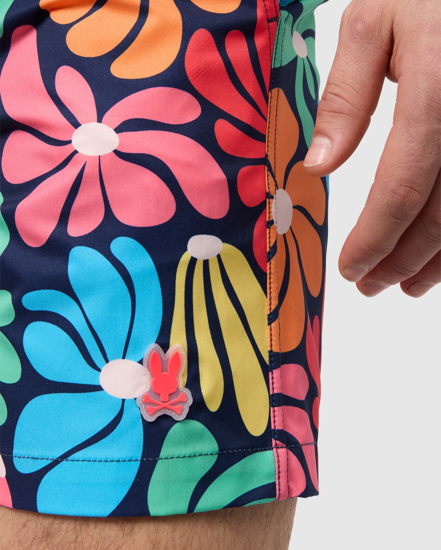 A close-up of a person's hand resting near their colorful quick-dry **Psycho Bunny MENS MENTZ SWIM TRUNK - B6W320B2SW** adorned with a vibrant flower pattern in pink, blue, green, yellow, and orange. There's a small pink bunny logo attached to the shorts near the hem.