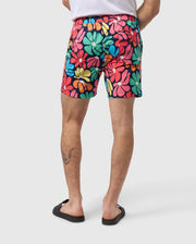 Back view of a person wearing MENS MENTZ SWIM TRUNK - B6W320B2SW by Psycho Bunny, standing with their hands by their sides. They are also wearing black flip-flops, and there is a visible tattoo on their left leg. The background is plain white.
