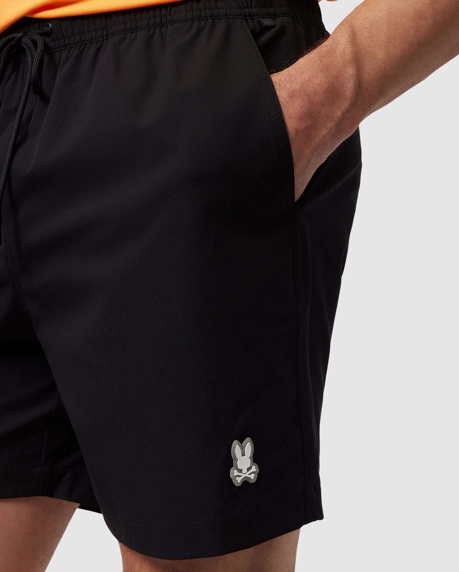 Close-up of a person wearing black athletic shorts with mechanical stretch and a small silver Psycho Bunny logo on the left thigh, detail of hand adjusting the drawstring waist.