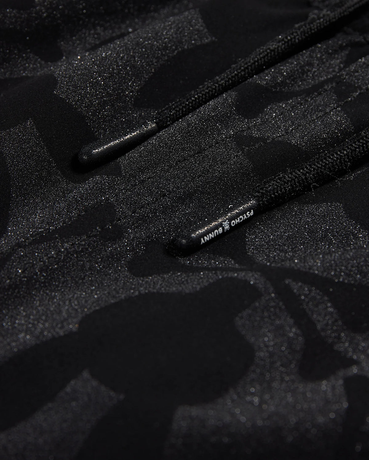Close-up of a textured black fabric with a subtle camo pattern and glittery specks, featuring a drawstring with visible branding on the Psycho Bunny MENS MALTA HYDROCHROMIC SWIM TRUNK - B6W321B2SW.
