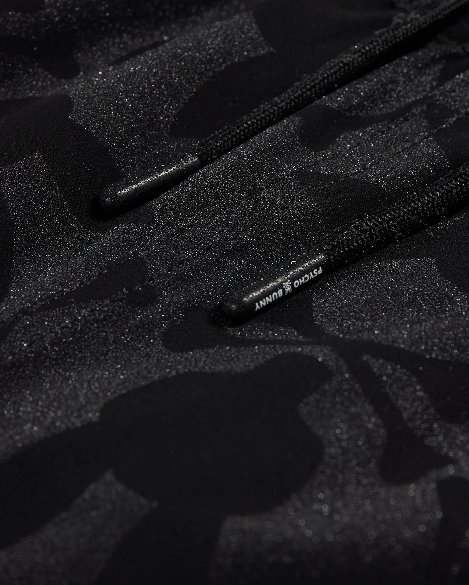 Close-up of a textured black fabric with a subtle camo pattern and glittery specks, featuring a drawstring with visible branding on the Psycho Bunny MENS MALTA HYDROCHROMIC SWIM TRUNK - B6W321B2SW.