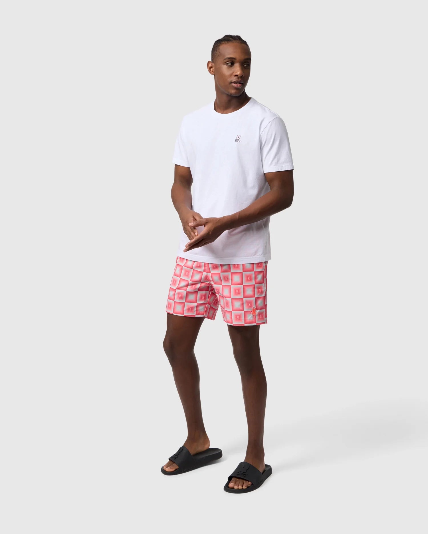 A man in a white t-shirt and red Psycho Bunny UTICA AOP swim trunk stands in a neutral stance against a light gray background, looking to the side. He wears black flip-flops.
