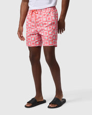 A person wears patterned pink and white Psycho Bunny AOP swim trunks and black slide sandals, shown from waist to calves with a plain white background.
