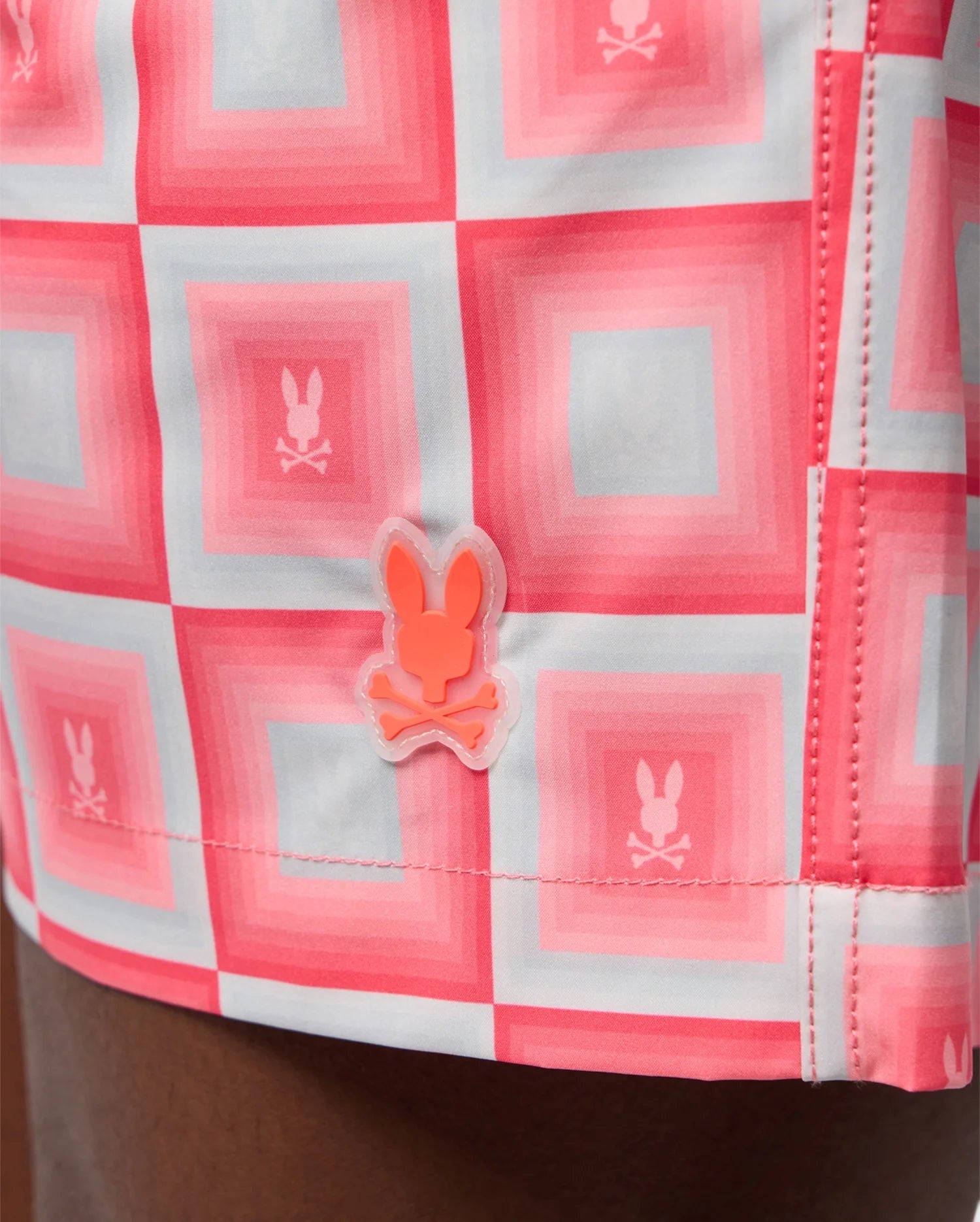 Close-up of a Psycho Bunny MENS UTICA SWIM TRUNK - B6W399B2SW with a red and pink grid pattern featuring repeated bunny logos, and a bunny-shaped pin attached to it.