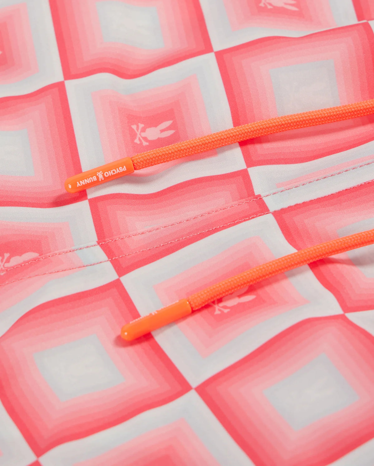 Close-up view of a Psycho Bunny orange shoelace on a pink and white checked fabric, with the text "running" embossed on the lace tip, depicting an all-over print style.