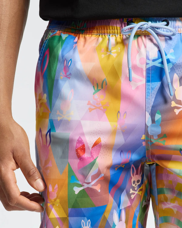 Close-up of a person wearing Psycho Bunny's MENS MILOS PRINTED SWIM TRUNK - B6W446F200, featuring colorful geometric patterns and playful bunny and skull motifs. Made from quick-dry fabric with a drawstring waist, perfect for vacation. The person pairs it with a black top, their left hand resting by their side.