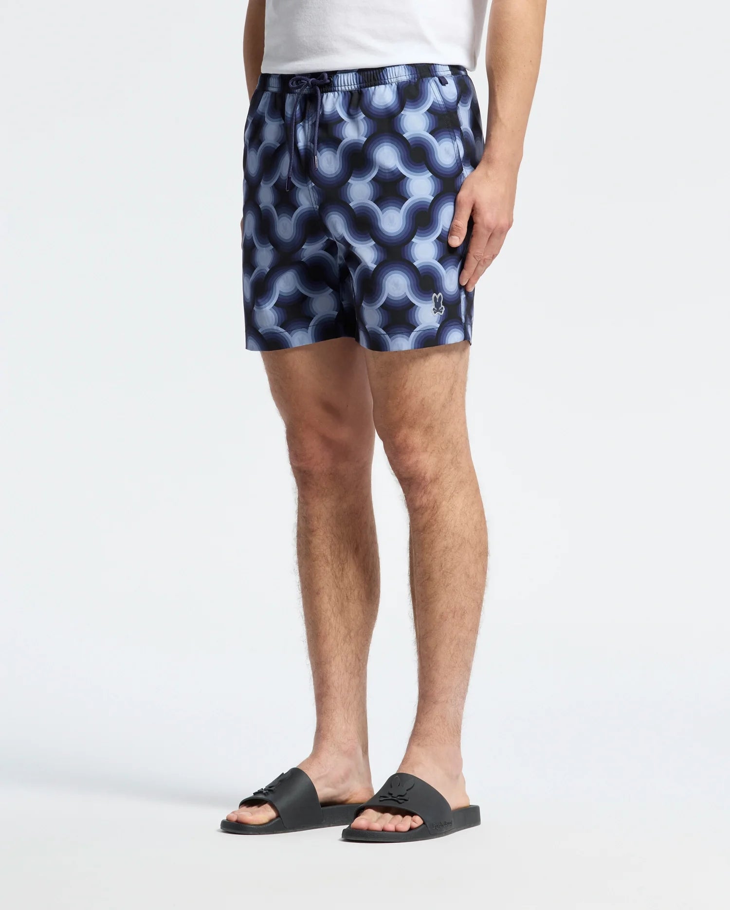 A person wearing Psycho Bunny Paros swim trunks, styled with a white T-shirt and black slides, stands against a plain background, only their lower half is visible.