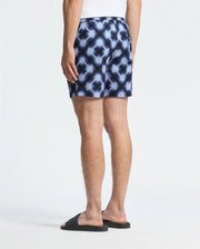 Rear view of a person wearing Psycho Bunny's MENS PAROS PRINTED SWIM TRUNK - B6W450F200 in blue and white geo-pattern, paired with black slide sandals against a plain white background.