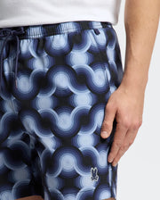 A man wears Psycho Bunny's MENS PAROS PRINTED SWIM TRUNK (B6W450F200) with a geo-patterned wave design and small bunny logo. His right hand rests next to the shorts, perfectly complementing his white shirt.