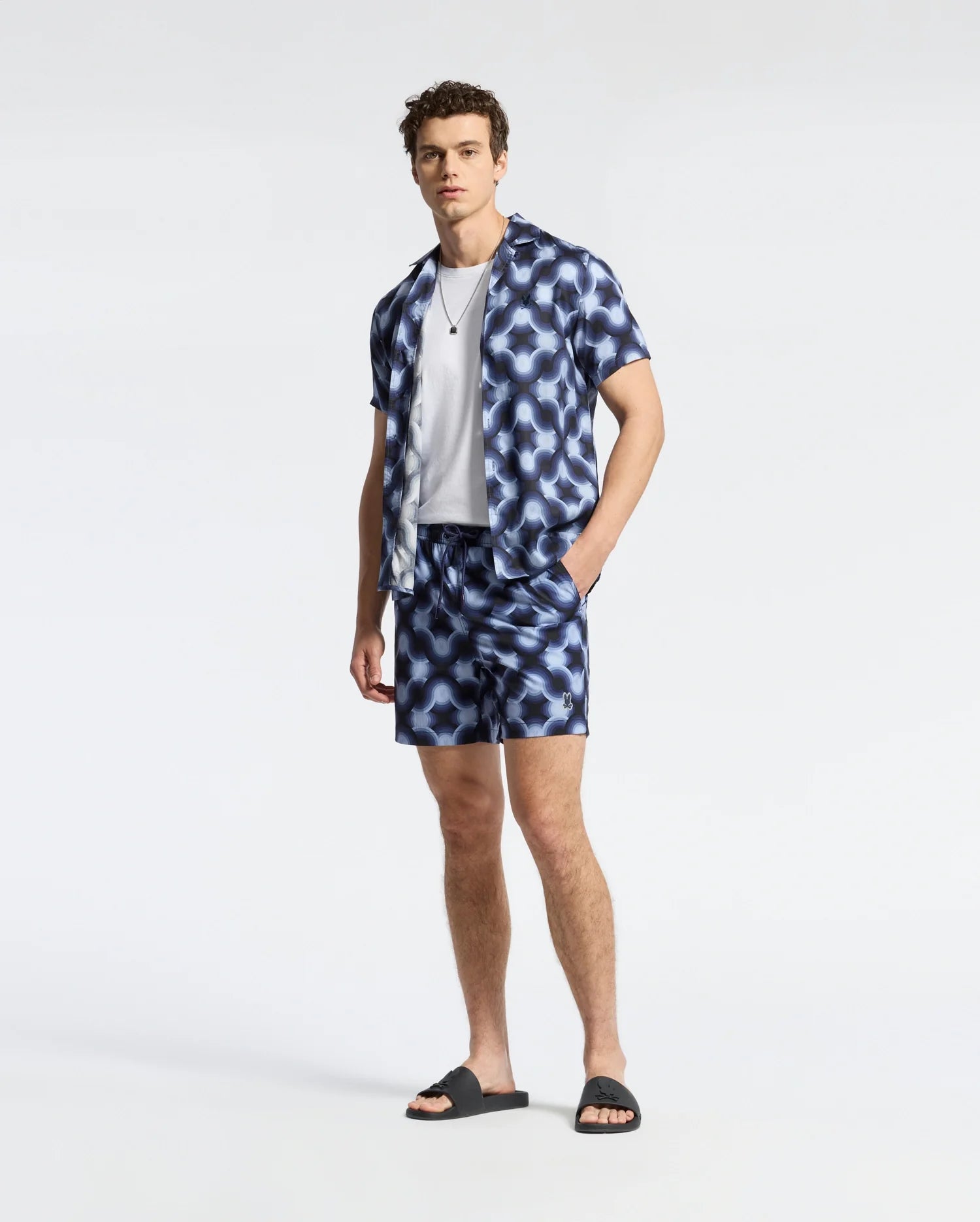 A person stands in Psycho Bunny's blue geo-patterned MENS PAROS PRINTED SWIM TRUNK over a white tee, paired with black slide sandals and a necklace, holding a waterproof bag against a plain light-colored background.
