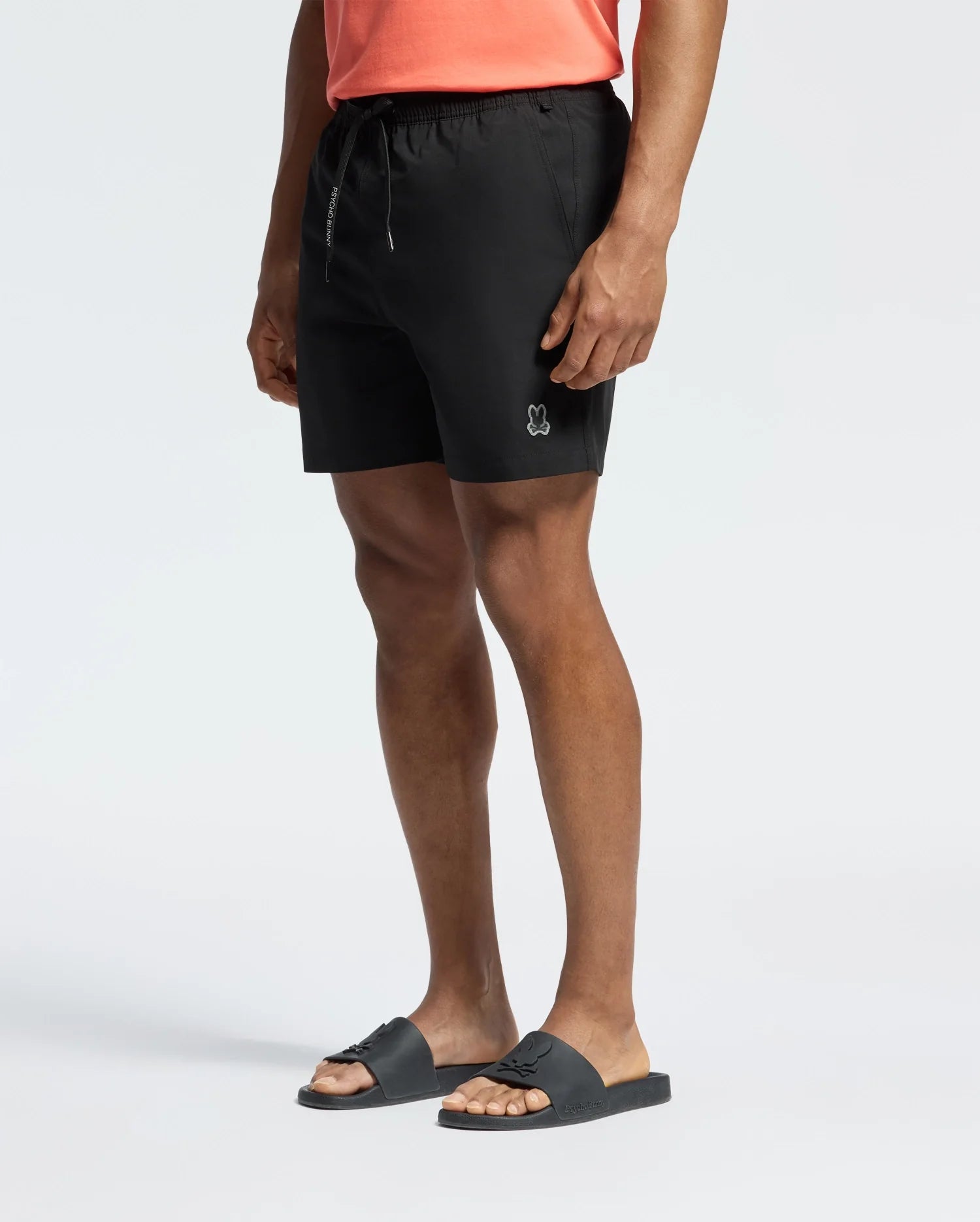 A person in a coral shirt and Psycho Bunny swim trunks (B6W453F200) with a small bunny logo stands with one hand in their pocket. They sport hydrochromic slides, ideal for any pool party, against a plain white background.