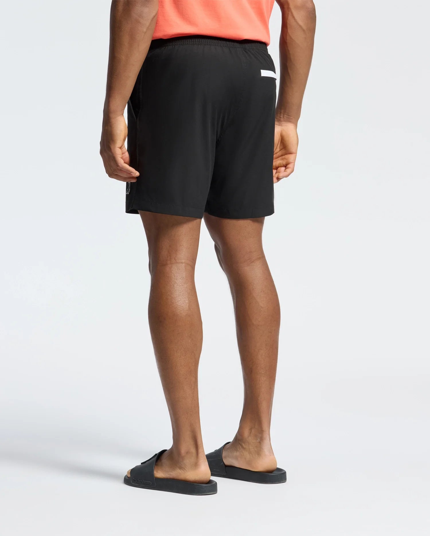 A person is wearing a coral T-shirt, Psycho Bunny black slide sandals, and MENS SAMOS HYDROCHROMIC PRINTED SWIM TRUNK - B6W453F200 by Psycho Bunny, which features a small white label on the back. The setting is a plain, light background, ideal for gearing up for a pool party.
