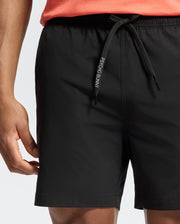 Someone is wearing Psycho Bunny's MENS SAMOS HYDROCHROMIC PRINTED SWIM TRUNK in black, showcasing the tailored fit and distinctive logo. Paired with a coral top, these shorts feature an elastic waistband and subtle hydrochromic detailing, making them ideal for a pool party.