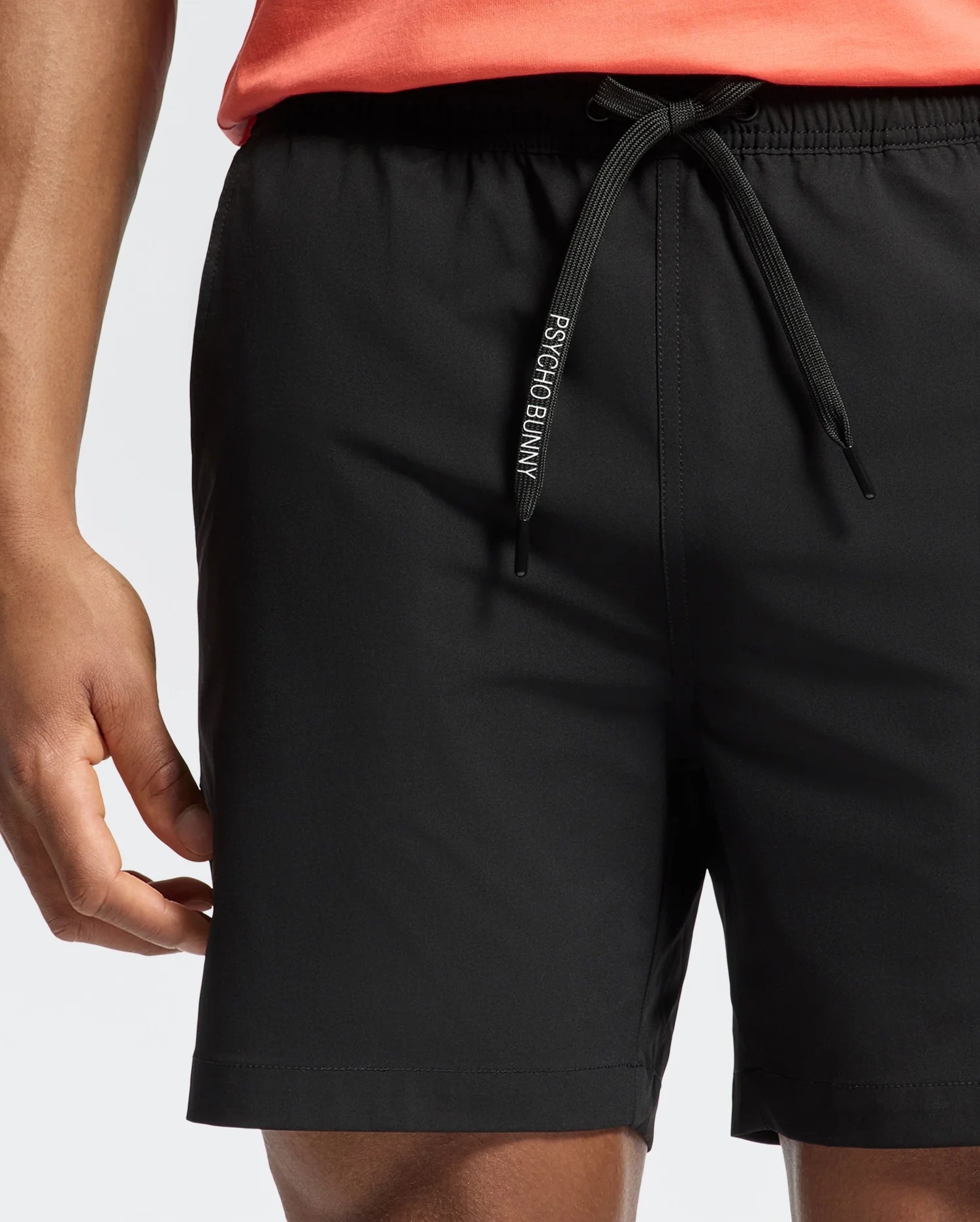 Someone is wearing Psycho Bunny's MENS SAMOS HYDROCHROMIC PRINTED SWIM TRUNK in black, showcasing the tailored fit and distinctive logo. Paired with a coral top, these shorts feature an elastic waistband and subtle hydrochromic detailing, making them ideal for a pool party.