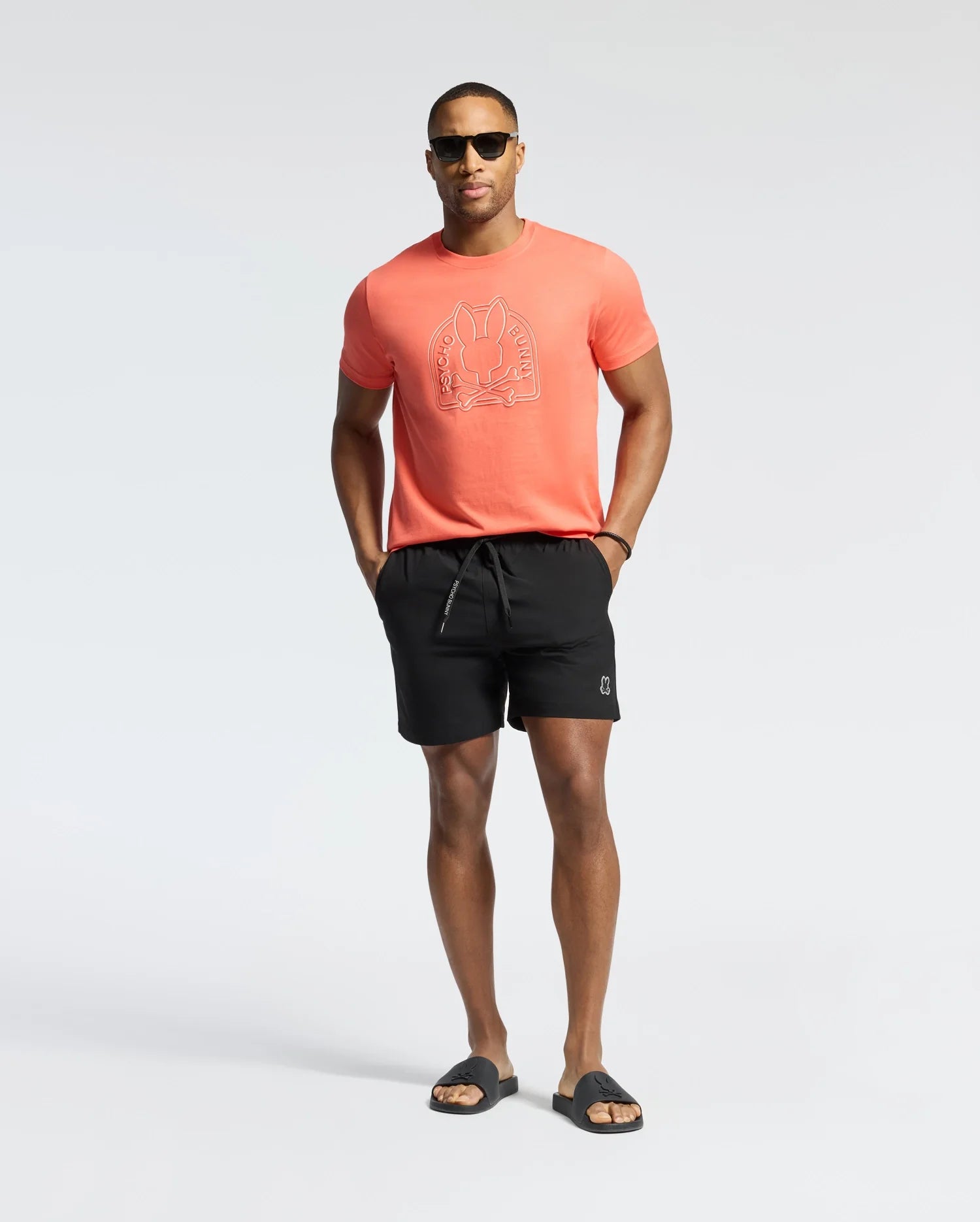 A man confidently poses against a plain white background, wearing sunglasses, Psycho Bunny's bright orange graphic T-shirt, MENS SAMOS HYDROCHROMIC PRINTED SWIM TRUNK - B6W453F200 in black, and black slide sandals, perfect for a pool party. Hands rest casually in pockets.