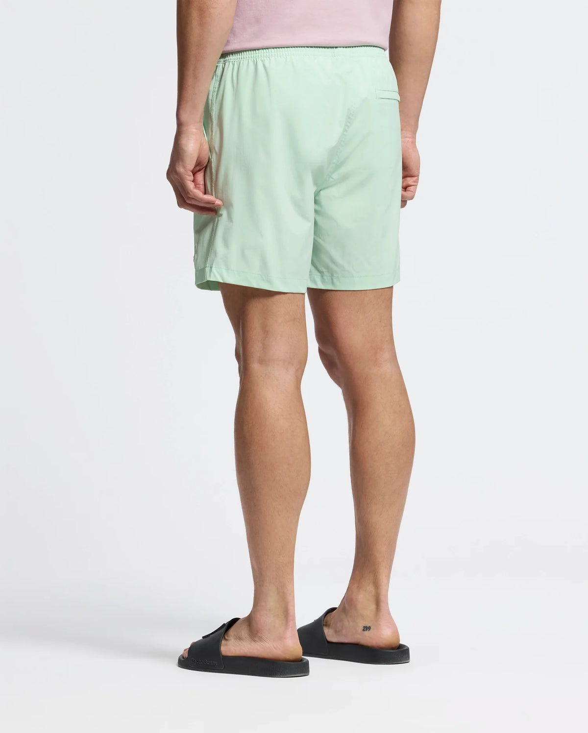 A person wearing Psycho Bunny's MENS SAMOS HYDROCHROMIC PRINTED SWIM TRUNK (B6W453F200) and black slide sandals stands with their back to the camera, against a plain white canvas.