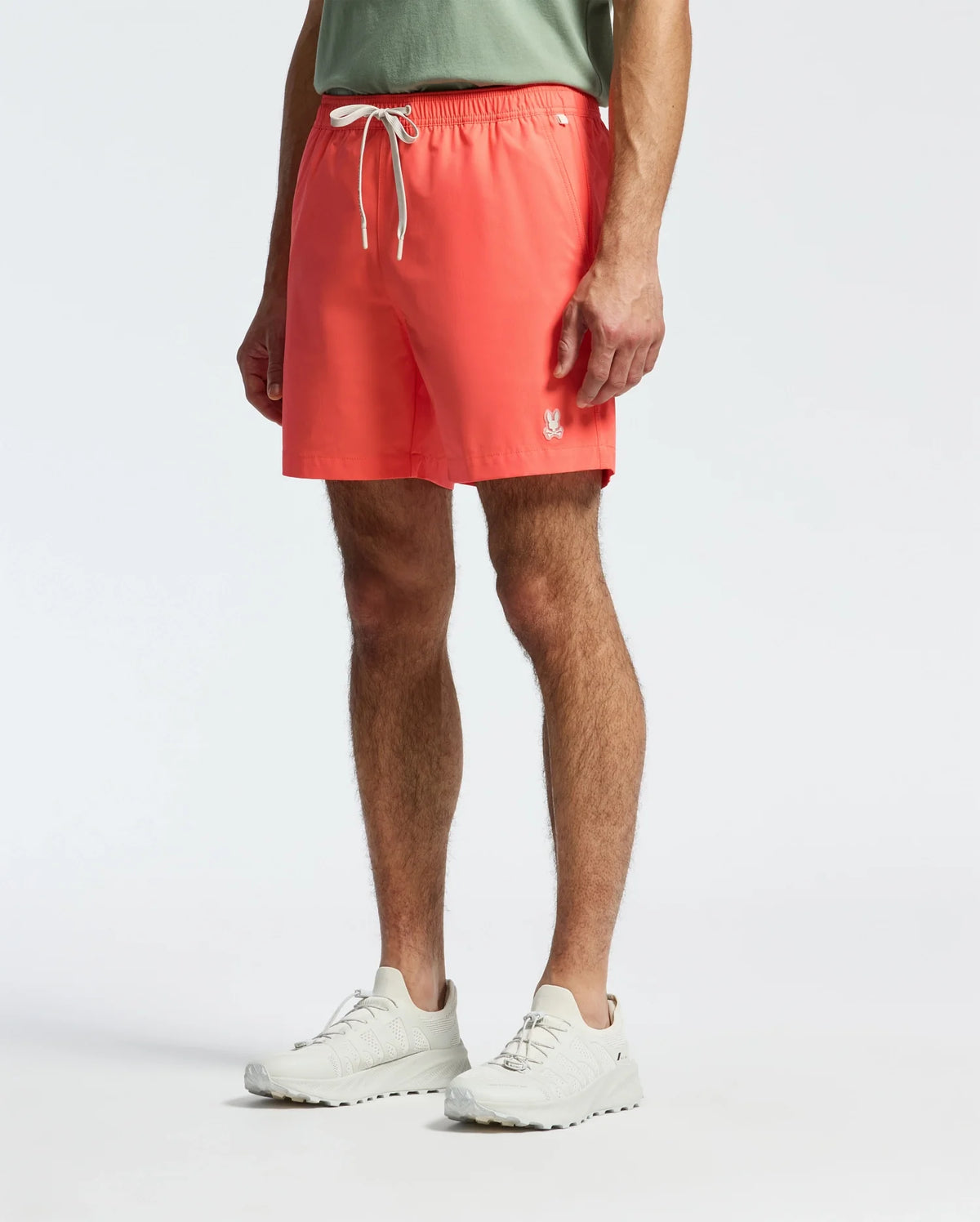 A person in a light green T-shirt and Psycho Bunny's MENS SAMOS HYDROCHROMIC SWIM TRUNK in bright coral with a small logo on the left leg stands against a plain backdrop, ready for a pool party. They complete their casual look with white athletic shoes.