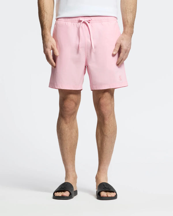 A person in light pink Psycho Bunny MENS SAMOS HYDROCHROMIC PRINTED SWIM TRUNKS, featuring innovative hydrochromic technology, stands against a plain white background wearing a white shirt and black slide sandals.