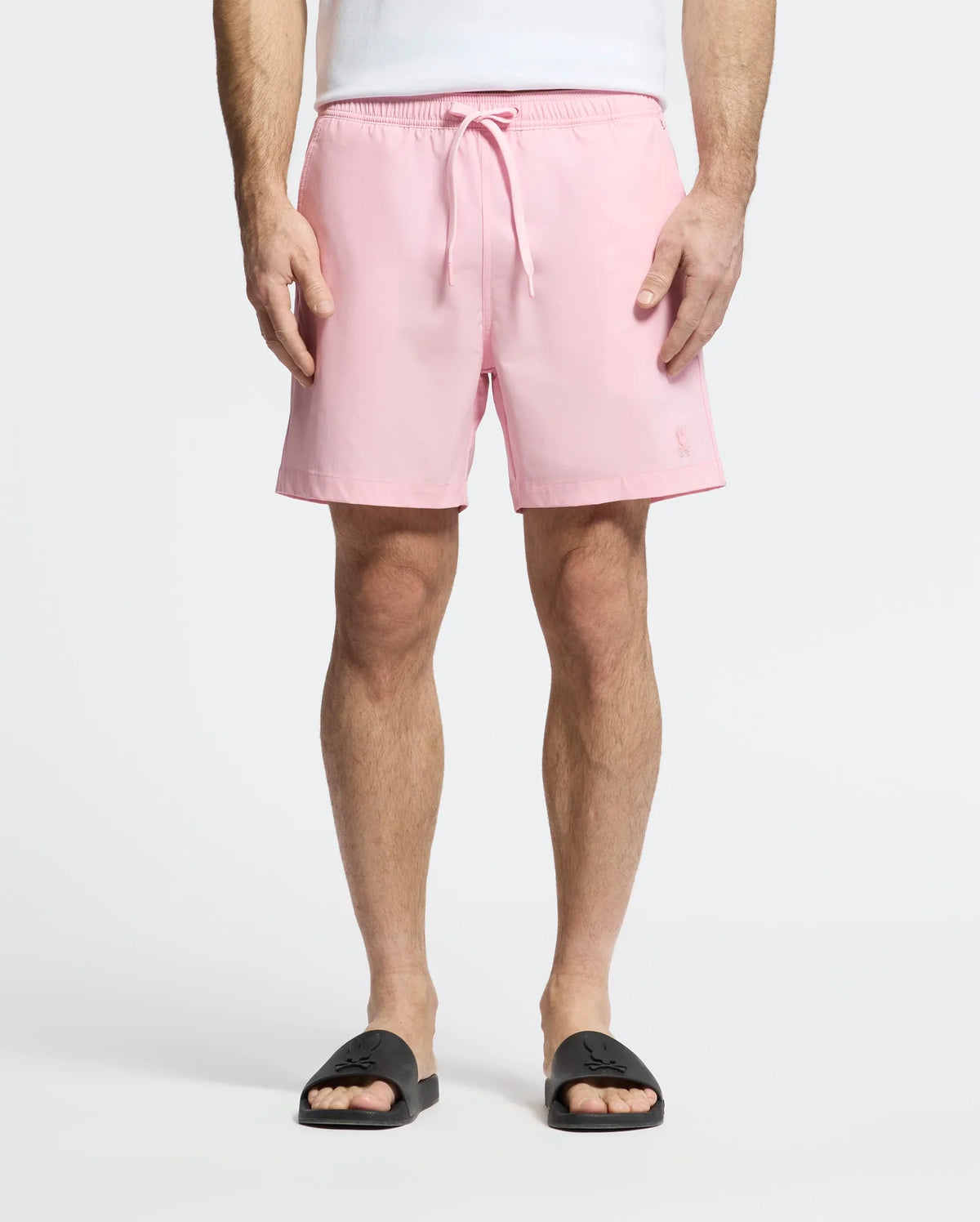 A person in light pink Psycho Bunny MENS SAMOS HYDROCHROMIC PRINTED SWIM TRUNKS, featuring innovative hydrochromic technology, stands against a plain white background wearing a white shirt and black slide sandals.
