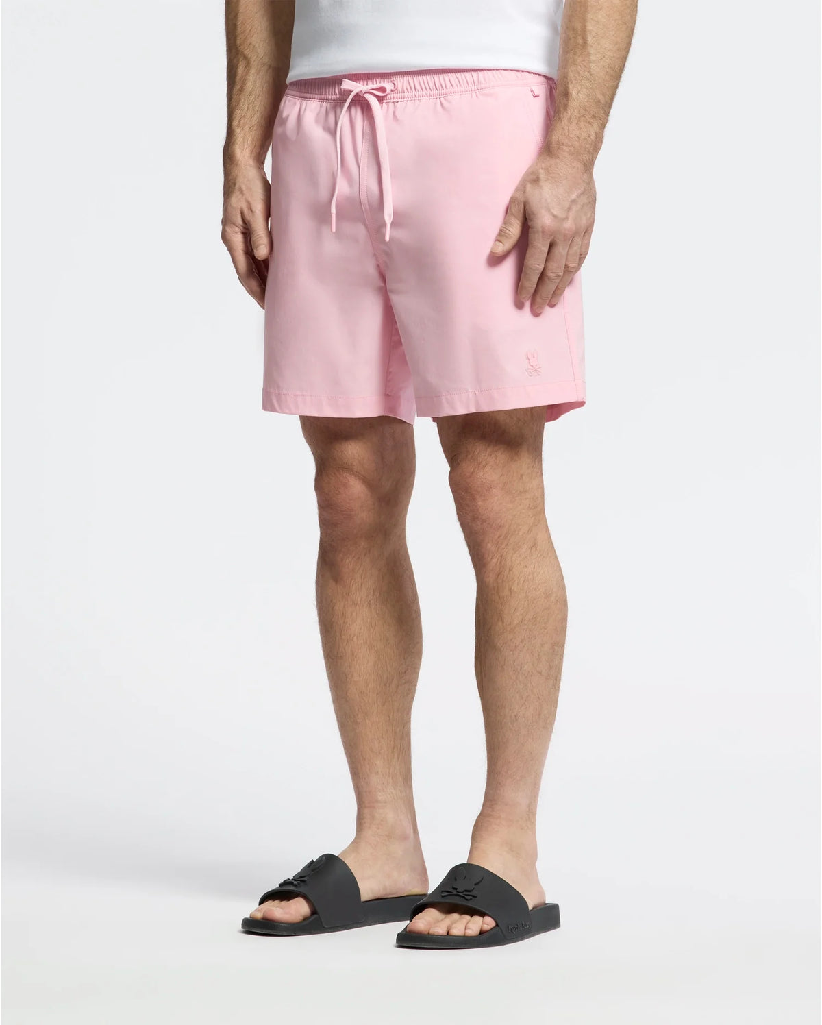 Wearing Psycho Bunny's MENS SAMOS HYDROCHROMIC PRINTED SWIM TRUNK in light pink, a person stands against a plain background. Paired with black slide sandals, the focus is on the comfortable, casual style created by the trunks' mesh lining and drawstring waist.
