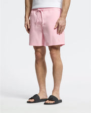 Wearing Psycho Bunny's MENS SAMOS HYDROCHROMIC PRINTED SWIM TRUNK in light pink, a person stands against a plain background. Paired with black slide sandals, the focus is on the comfortable, casual style created by the trunks' mesh lining and drawstring waist.