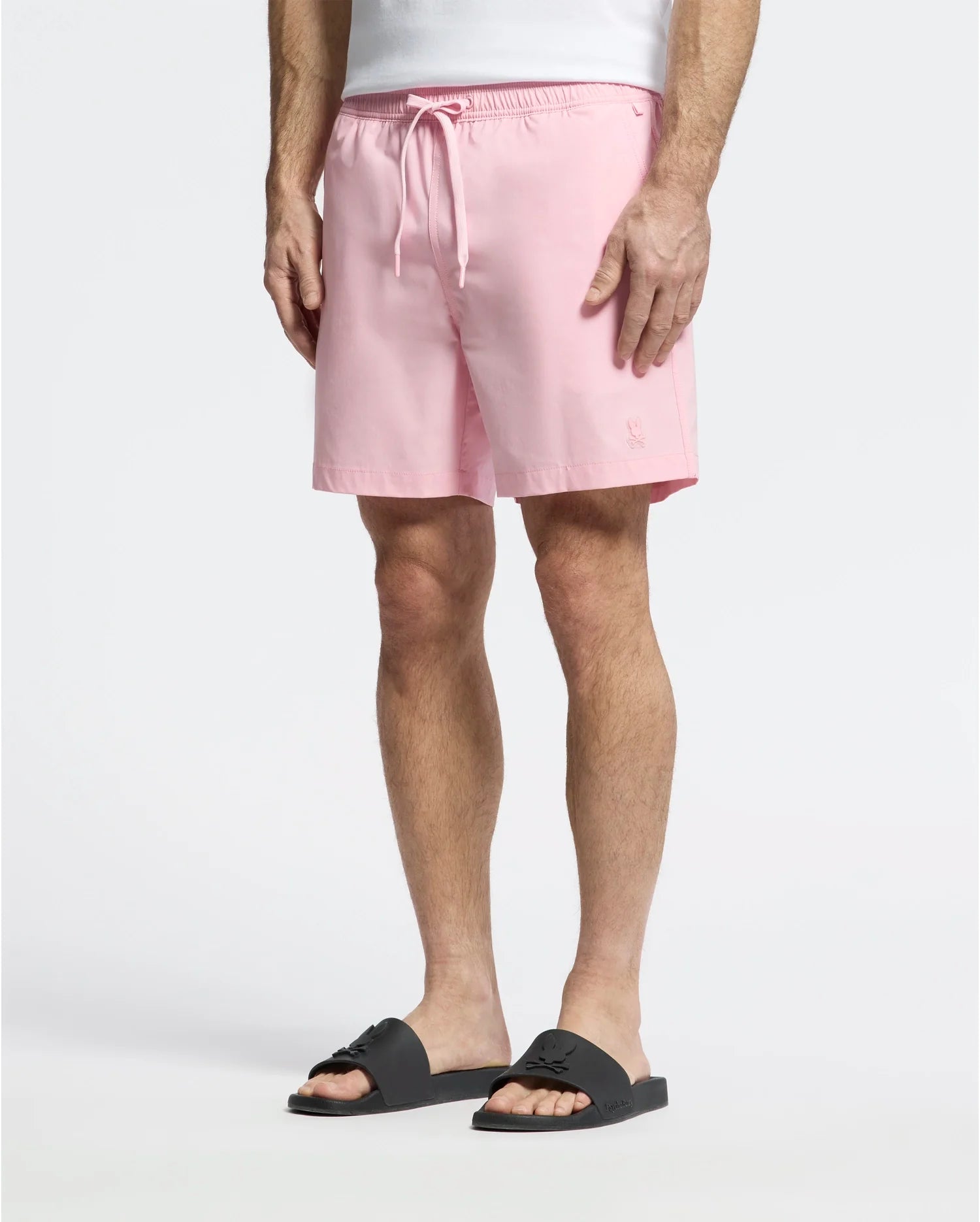 Wearing Psycho Bunny's MENS SAMOS HYDROCHROMIC PRINTED SWIM TRUNK in light pink, a person stands against a plain background. Paired with black slide sandals, the focus is on the comfortable, casual style created by the trunks' mesh lining and drawstring waist.