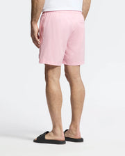 A person stands in Psycho Bunny's Samos Hydrochromic swim trunks, a white top, and black slide sandals against a plain white backdrop, showcasing their casual attire from behind.