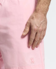 A person wears the Psycho Bunny MENS SAMOS HYDROCHROMIC PRINTED SWIM TRUNK in pink, featuring a small bunny logo. Their right hand rests near their leg. These trunks use hydrochromic technology that changes color when wet, creating a vibrant background against the plain white.