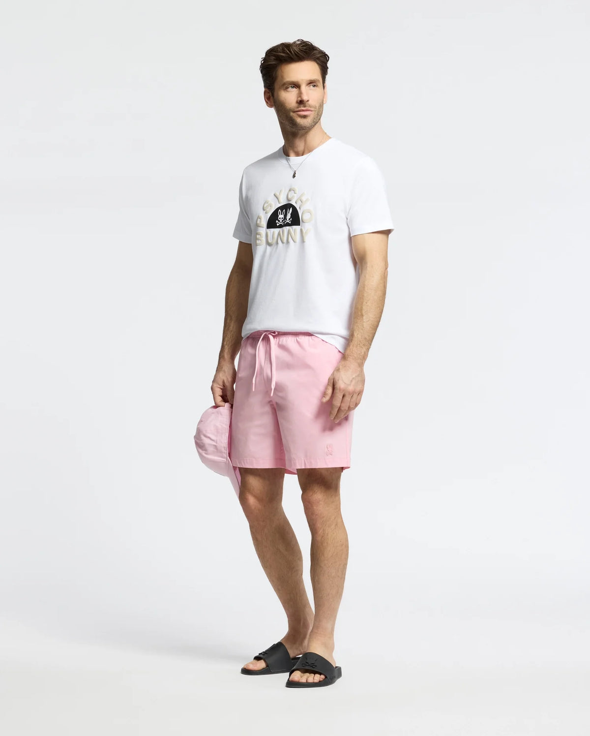 A man in a white graphic t-shirt and Psycho Bunny MENS SAMOS HYDROCHROMIC PRINTED SWIM TRUNK, paired with black slide sandals and holding a matching pink cap, gazes off to the side against a plain white background.