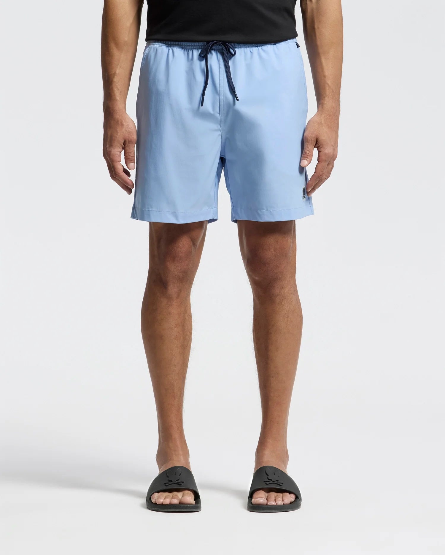 A man in Psycho Bunny's MENS SAMOS HYDROCHROMIC PRINTED SWIM TRUNK - B6W453F200 stands against a white background, paired with a black shirt and sandals. His relaxed arms amplify the casual vibe of his ensemble.