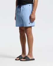 A person in Psycho Bunny's light blue MENS SAMOS HYDROCHROMIC PRINTED SWIM TRUNK - B6W453F200 with an emblem on the leg stands against a plain background, wearing black sandals and a sleek black top effortlessly.