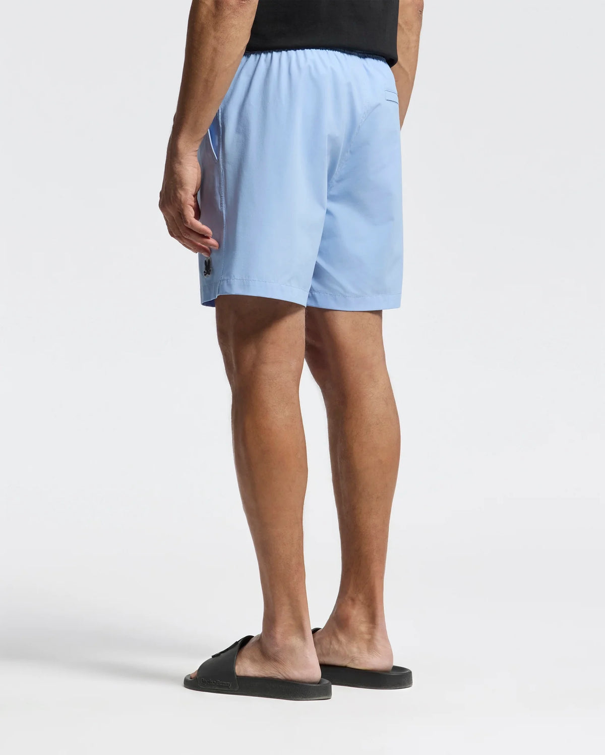 A person in Psycho Bunny's light blue MENS SAMOS HYDROCHROMIC PRINTED SWIM TRUNK - B6W453F200 with an elastic waistband and black slides stands against a white backdrop. The trunks subtly change hues when wet, and the individual faces away with their left arm hanging loosely by their side.