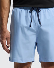 A person is wearing Psycho Bunny's MENS SAMOS HYDROCHROMIC PRINTED SWIM TRUNK in light blue with a dark blue drawstring. The image highlights the midsection to thighs, emphasizing the trunks' color and unique hydrochromic technology.