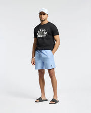 In a studio, a man sports a black Psycho Bunny T-shirt, MENS SAMOS Hydrochromic printed swim trunks in light blue, black slide sandals, and a white cap. With his relaxed pose against the white backdrop and his waterproof bag nearby, he's set for any adventure.