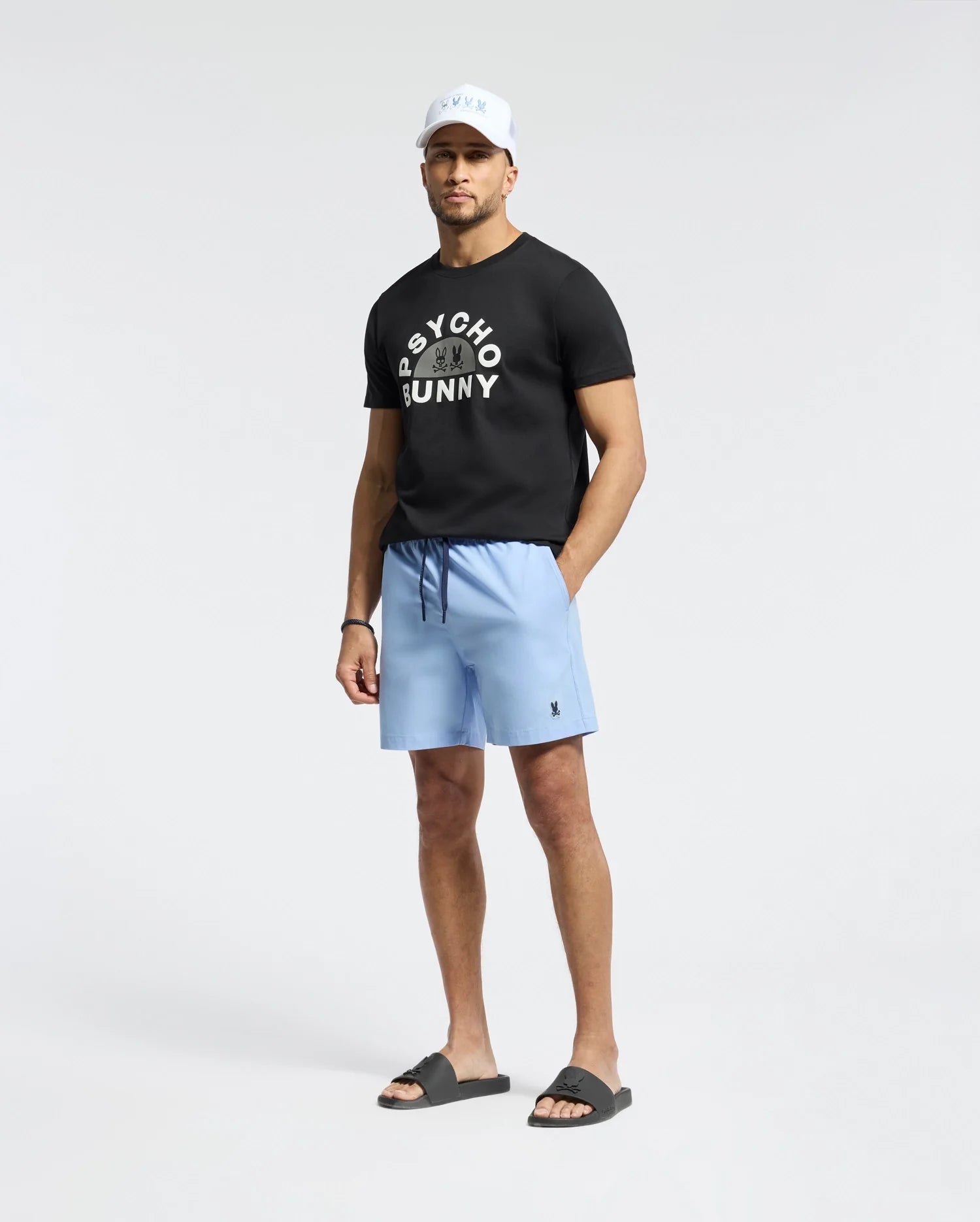 In a studio, a man sports a black Psycho Bunny T-shirt, MENS SAMOS Hydrochromic printed swim trunks in light blue, black slide sandals, and a white cap. With his relaxed pose against the white backdrop and his waterproof bag nearby, he's set for any adventure.