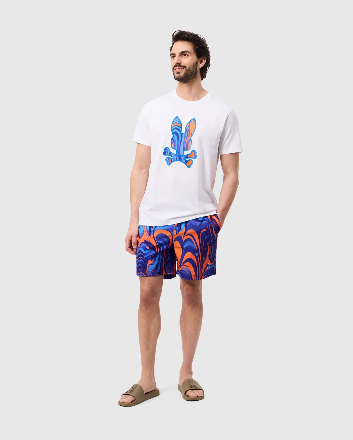 A man stands against a plain backdrop, wearing a white T-shirt with a colorful abstract design on the front and vibrant Psycho Bunny MENS NEVADA ALL OVER PRINT SWIM TRUNK - B6W526C200 made of quick-dry fabric. He is also wearing beige slide sandals. His left hand is in his pocket, and he is looking slightly to the side.