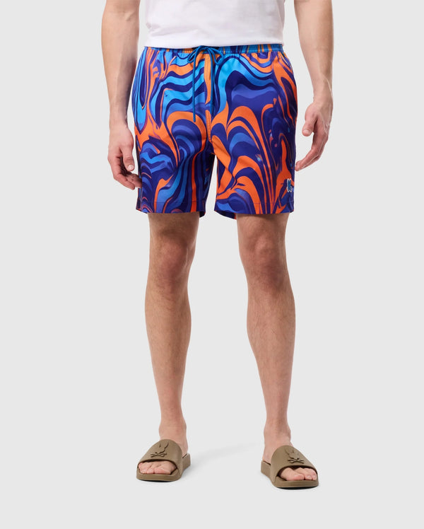A person wearing bright, multi-colored MENS NEVADA ALL OVER PRINT SWIM TRUNK - B6W526C200 by Psycho Bunny featuring a vibrant swirl print in blue, orange, and purple. They are also wearing brown slide sandals and a white T-shirt. The background is plain white. The photo only shows the person from the waist down.