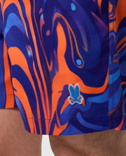 Close-up of a person wearing Psycho Bunny MENS NEVADA ALL OVER PRINT SWIM TRUNK - B6W526C200 featuring an abstract swirl print in orange and blue hues. Made from quick-dry fabric, the design showcases swirling lines and shapes. A small emblem of a stylized rabbit skull and crossbones is visible near the hem.