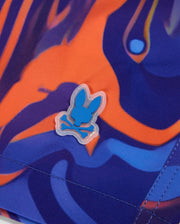 Close-up of a colorful fabric featuring a blend of orange, blue, and purple abstract patterns. The image also shows a small rubber emblem of a bunny skull and crossbones in blue and white, affixed to the vibrant swirl print of Psycho Bunny MENS NEVADA ALL OVER PRINT SWIM TRUNK - B6W526C200.