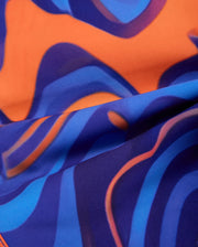 A close-up view of a fabric showing an abstract, wavy pattern with vibrant swirl print. The design predominantly features shades of blue and orange, creating a dynamic and visually engaging effect. The quick-dry fabric is smooth with gentle folds, making it perfect for the Psycho Bunny MENS NEVADA ALL OVER PRINT SWIM TRUNK - B6W526C200.