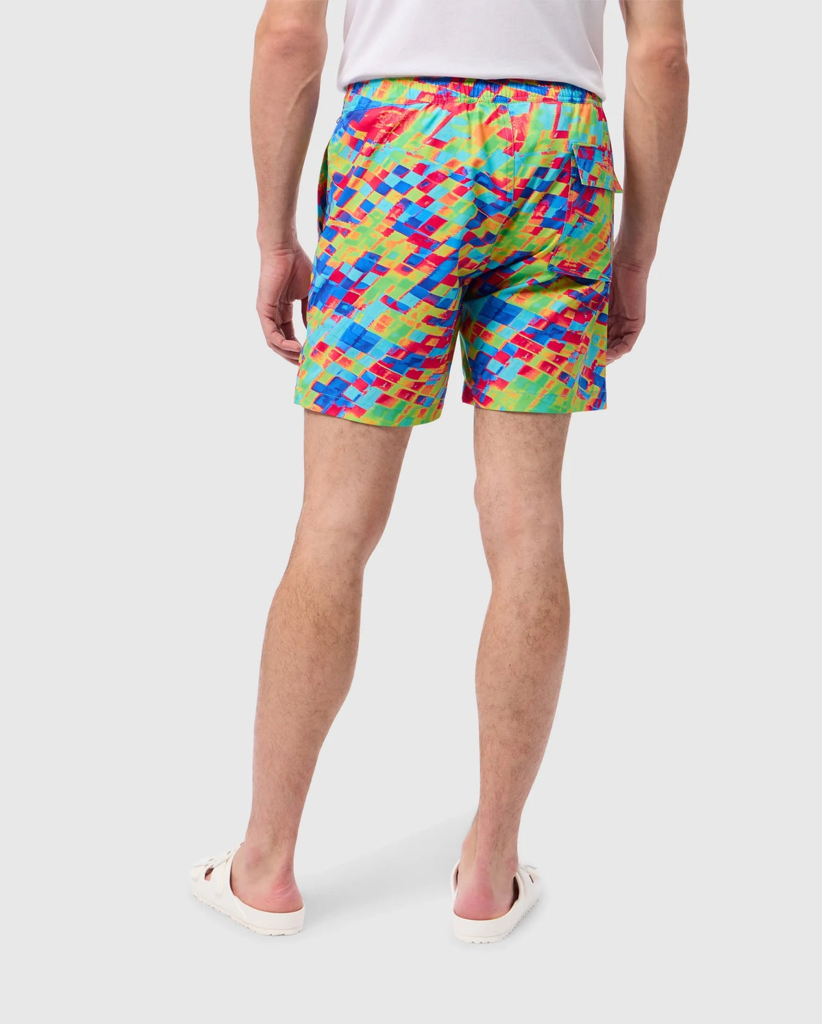 Back view of a person wearing colorful, patterned swim trunks named "MENS DENNIS ALL OVER PRINT SWIM TRUNK - B6W530C200" by Psycho Bunny, paired with a white shirt and white slide sandals. The shorts, crafted from quick-dry fabric, boast a vibrant mosaic-like design in shades of red, blue, yellow, and green. The person stands against a plain, light background.