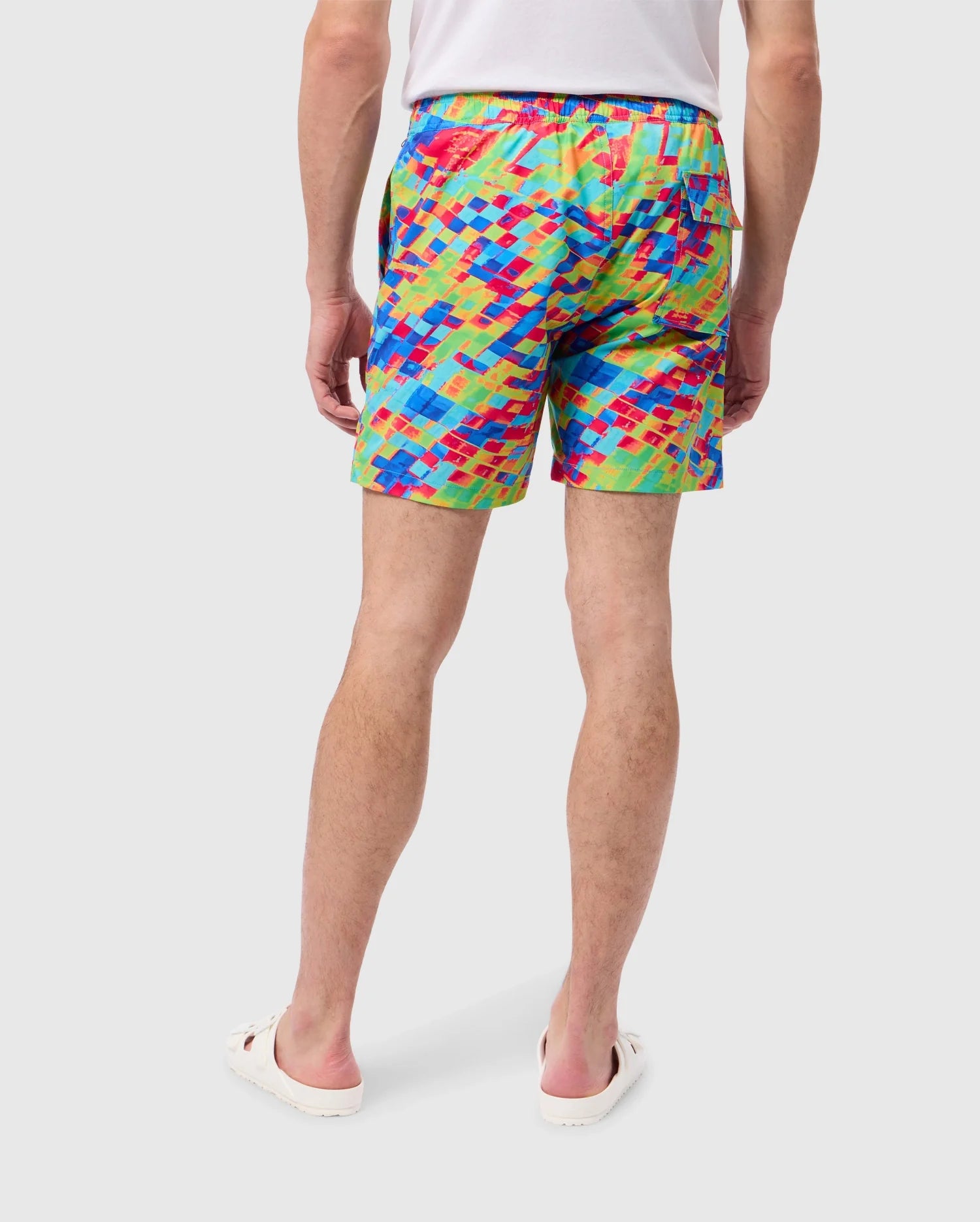 Back view of a person wearing colorful, patterned swim trunks named "MENS DENNIS ALL OVER PRINT SWIM TRUNK - B6W530C200" by Psycho Bunny, paired with a white shirt and white slide sandals. The shorts, crafted from quick-dry fabric, boast a vibrant mosaic-like design in shades of red, blue, yellow, and green. The person stands against a plain, light background.