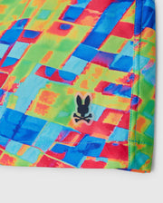 A close-up of the vibrant, multicolored MENS DENNIS ALL OVER PRINT SWIM TRUNK by Psycho Bunny, showcasing overlapping geometric shapes in red, blue, green, and yellow. The quick-dry fabric features a small black logo of a rabbit head over crossed bones. The stitching along the edge is visible.