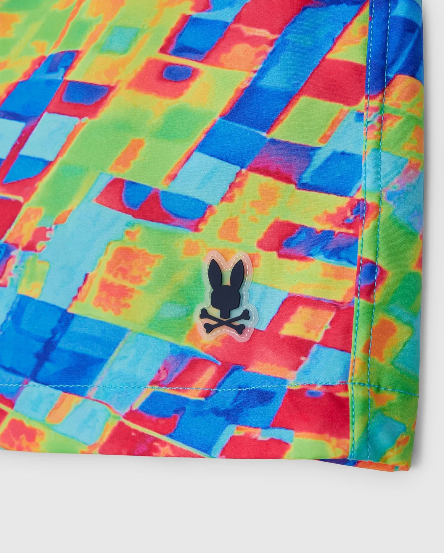 A close-up of the vibrant, multicolored MENS DENNIS ALL OVER PRINT SWIM TRUNK by Psycho Bunny, showcasing overlapping geometric shapes in red, blue, green, and yellow. The quick-dry fabric features a small black logo of a rabbit head over crossed bones. The stitching along the edge is visible.