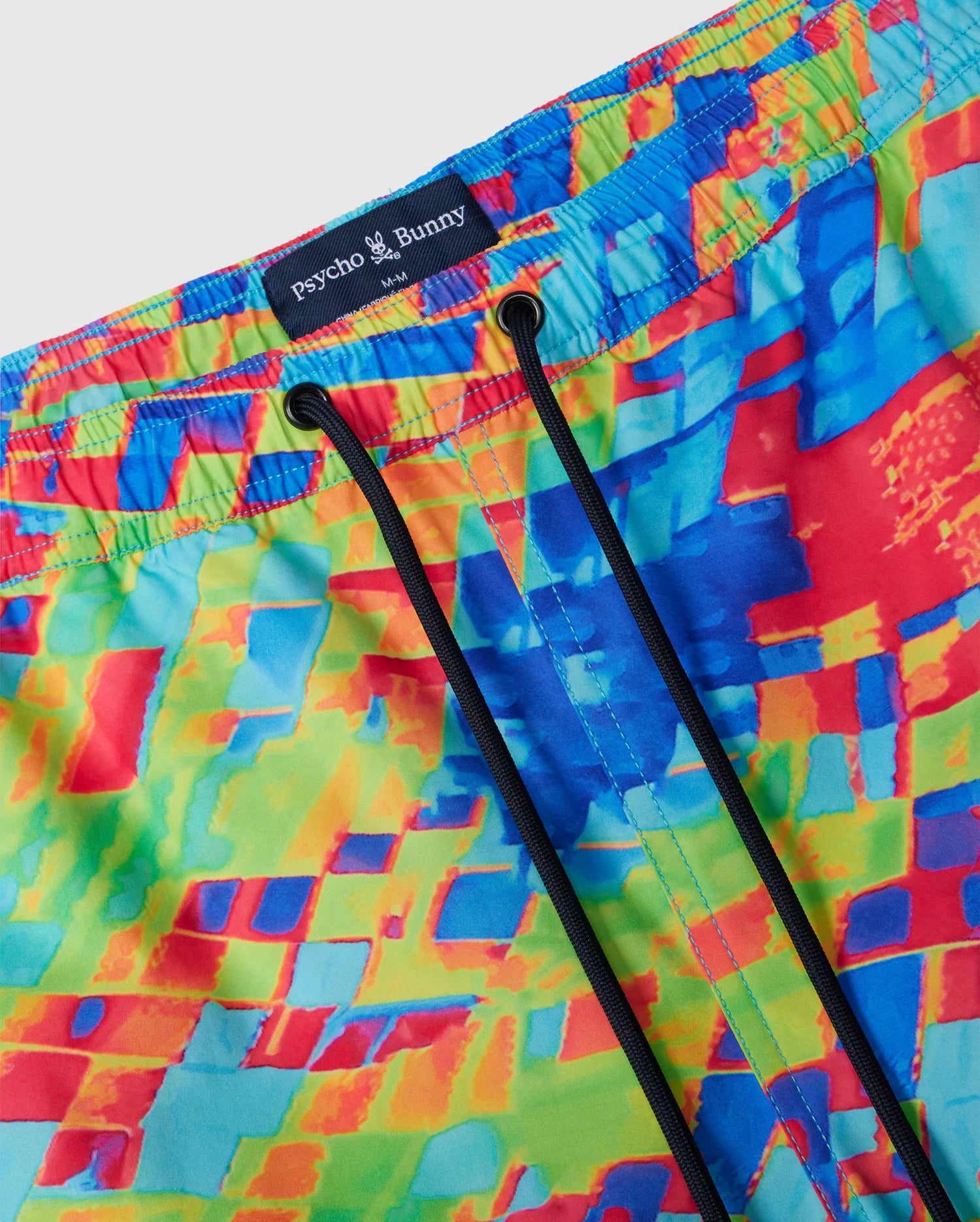 Close-up image of the MENS DENNIS ALL OVER PRINT SWIM TRUNK - B6W530C200 by Psycho Bunny, showcasing colorful swim trunks with an abstract, multicolored mosaic pattern in blue, red, yellow, and green. Made from quick-dry fabric, the waistband is elastic with black drawstrings. A tag at the waistband reads "Psycho Bunny" with a bunny logo.