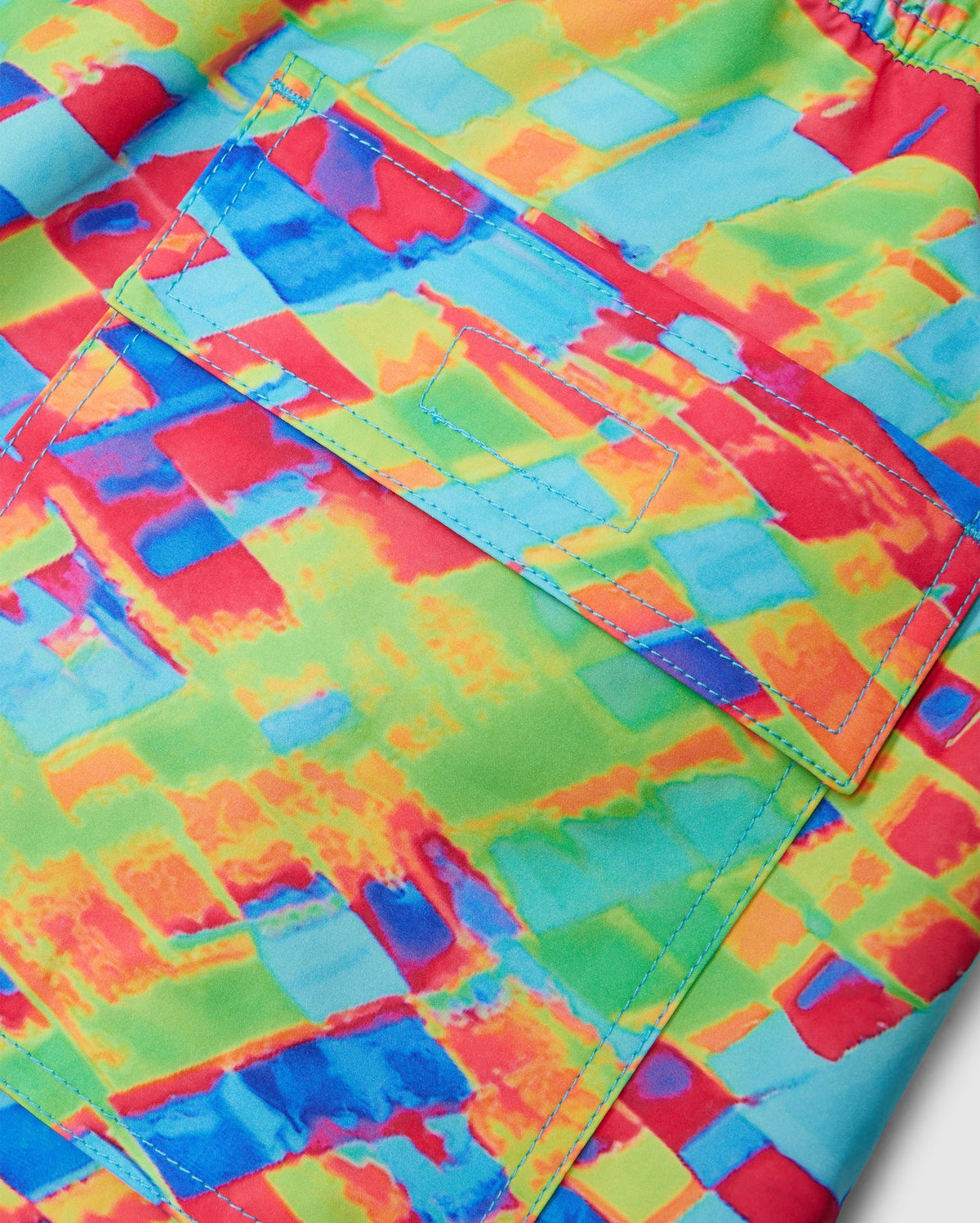 Close-up of a fabric showcasing the vibrant, multicolored pattern of the MENS DENNIS ALL OVER PRINT SWIM TRUNK - B6W530C200 by Psycho Bunny. The quick-dry material features shades of red, blue, green, and yellow, with an abstract and colorful design reminiscent of stylish swim trunks. A rectangular pocket with a flap is highlighted by detailed stitching in blue thread.