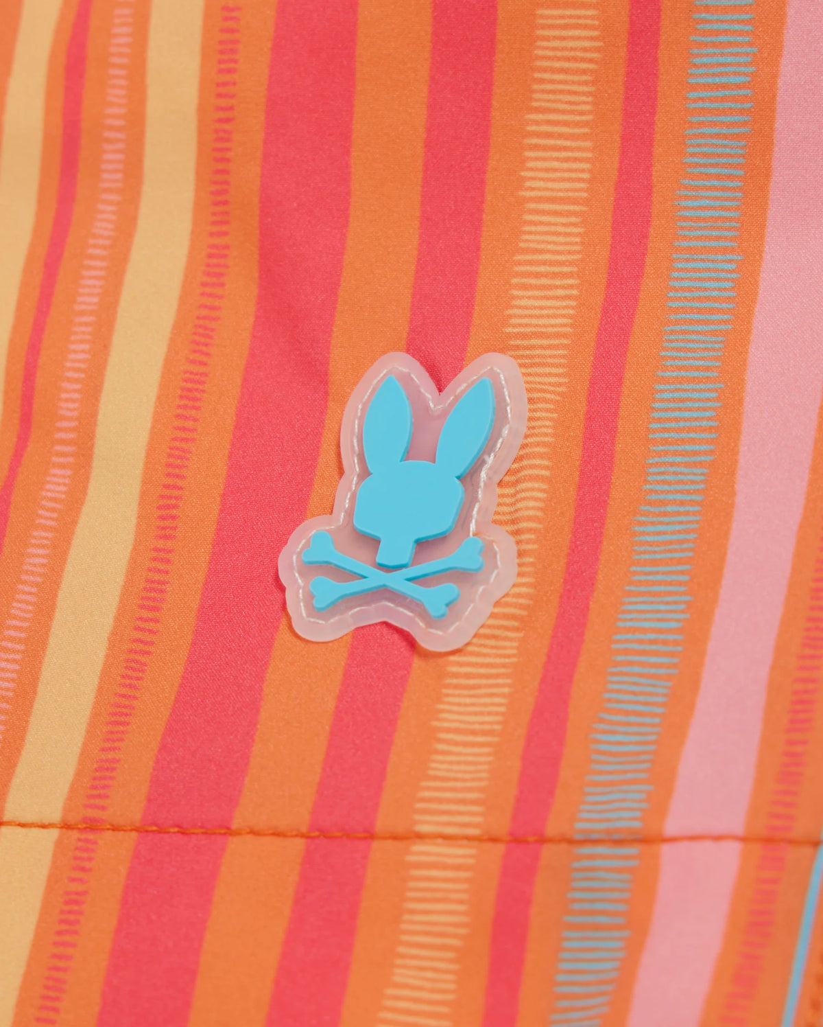 A colorful fabric displaying vertical stripes in shades of orange, pink, red, and yellow. An aqua blue bunny head with a crossbones symbol appears in the center, giving a playful and quirky touch to these vibrant quick-dry Psycho Bunny MENS ANTON ALL OVER PRINT SWIM TRUNK - B6W542C200.