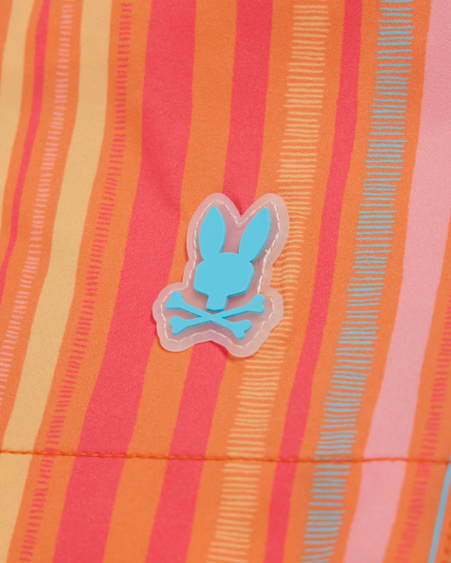 A colorful fabric displaying vertical stripes in shades of orange, pink, red, and yellow. An aqua blue bunny head with a crossbones symbol appears in the center, giving a playful and quirky touch to these vibrant quick-dry Psycho Bunny MENS ANTON ALL OVER PRINT SWIM TRUNK - B6W542C200.