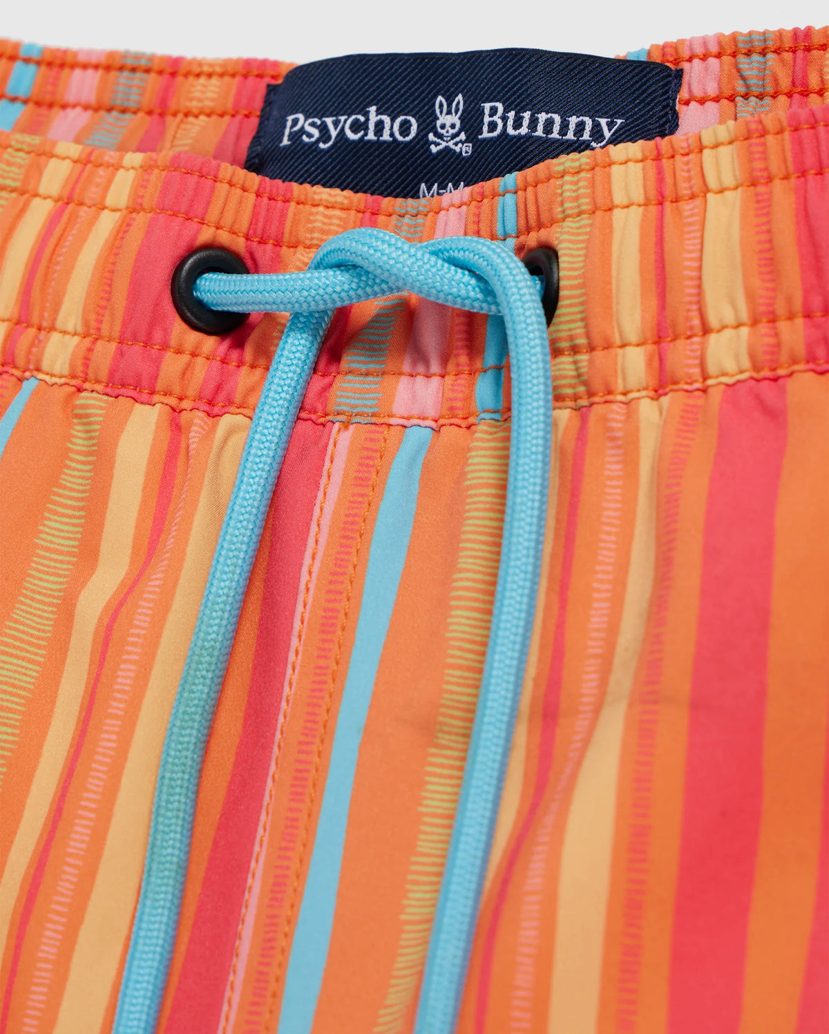 Close-up of colorful swim trunks with vertical orange, yellow, and blue stripes. The quick-dry waistband features a blue drawstring and a visible label with the brand name "Psycho Bunny," a small bunny logo, and the size "M." Product Name: MENS ANTON ALL OVER PRINT SWIM TRUNK - B6W542C200. Brand Name: Psycho Bunny.
